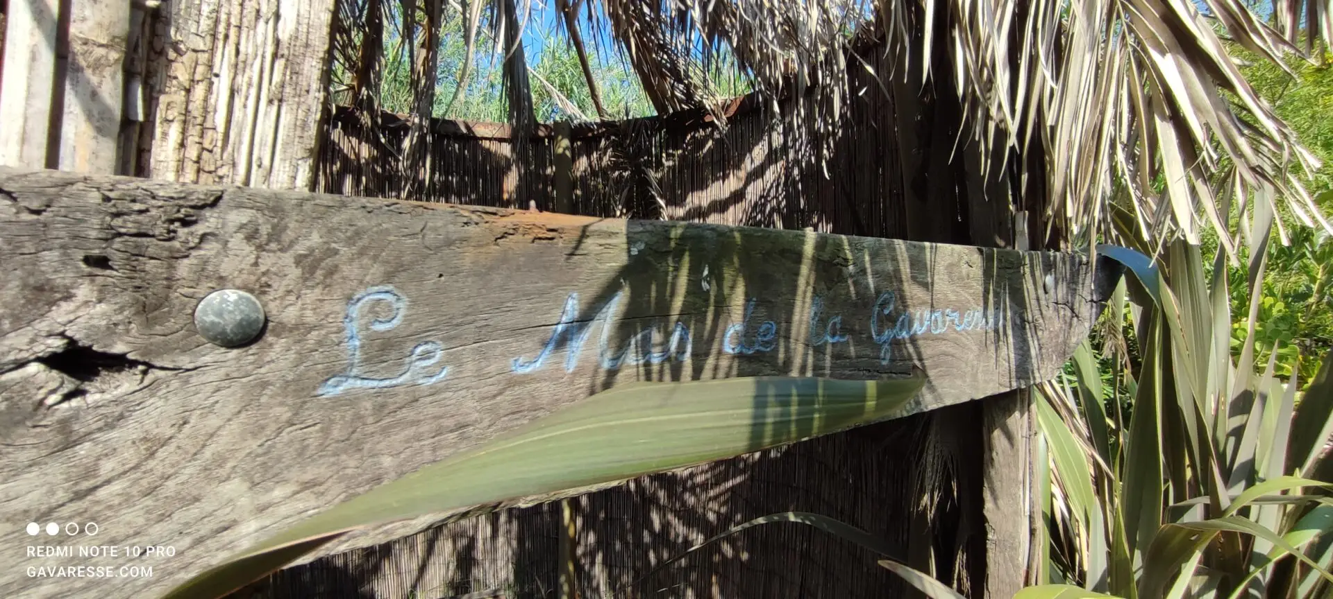 Gros plan sur une enseigne en bois "Le Mas de la Gavaresse" gravée sur un vieux morceau de tonneau, fixée à l'entrée d'une cabane d'enfant recouverte de roseaux.